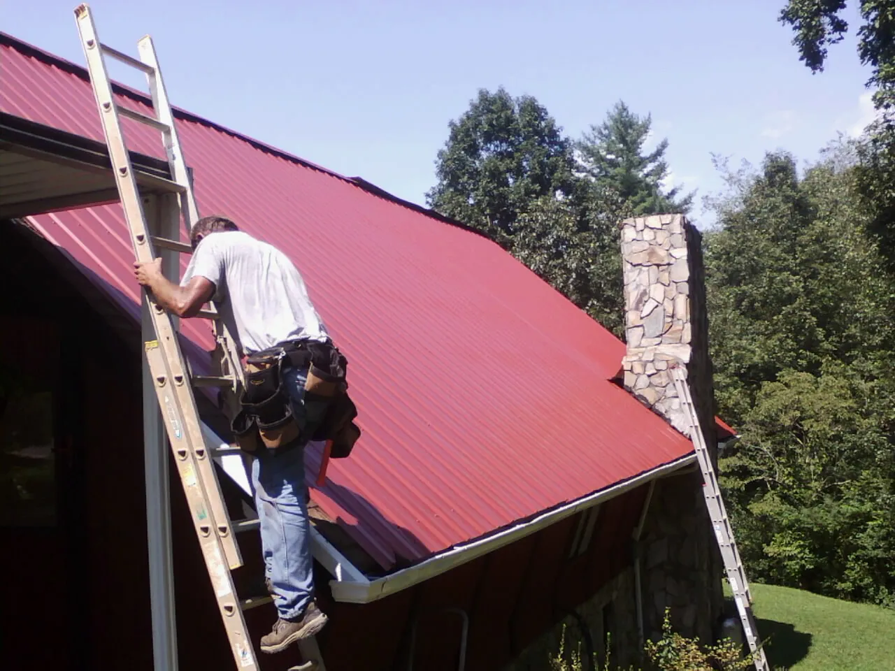 Roofer on a ladder beside a red metal roof and stone chimney — Soffit & Fascia Repair by Roofing Brothers in Gerton